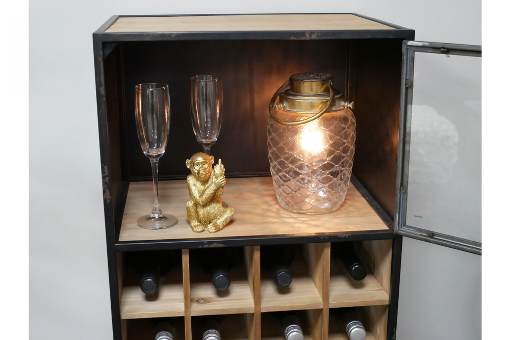 Black metal and light wood wine cabinet with glass door, holding bottles, two clear glasses, gold monkey figurine, and lit