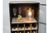Black metal and light wood wine cabinet with glass door, holding bottles, two clear glasses, gold monkey figurine, and lit