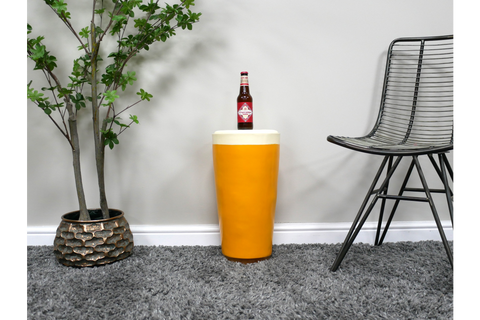 Round yellow side table with white top, ceramic material, shown with bottle on top, next to black wire chair and potted