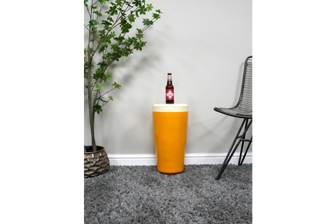 Cylindrical mustard yellow Lager Side Table with cream top, displayed on grey carpet between plant and black wire chair.