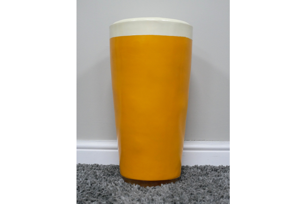 Lager Side Table shaped like a yellow and white pint glass, ceramic material, front view on grey carpet against white wall.