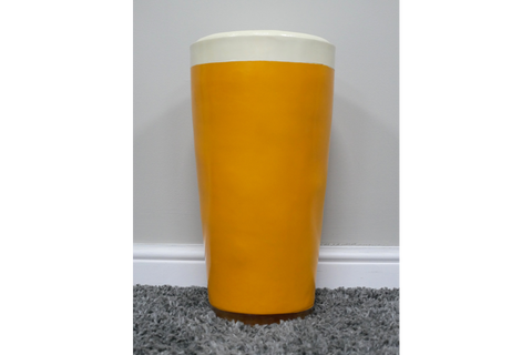 Lager Side Table shaped like a yellow and white pint glass, ceramic material, front view on grey carpet against white wall.