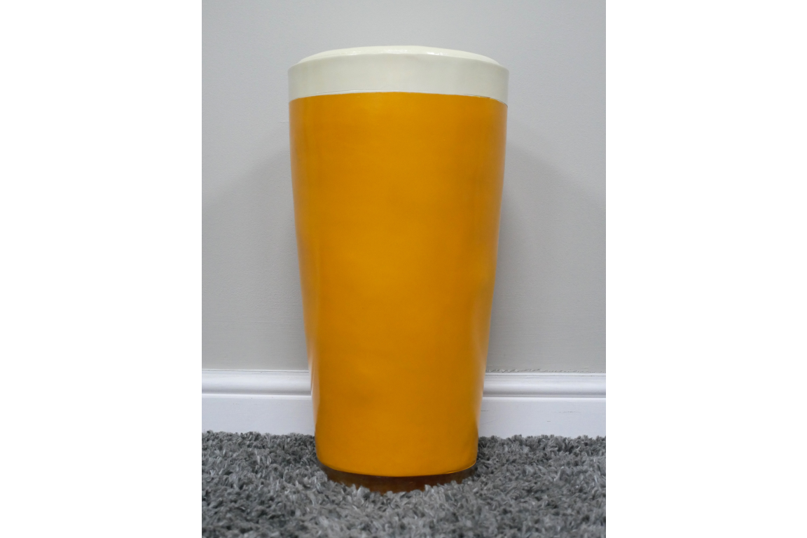 Lager Side Table shaped like a yellow and white pint glass, ceramic material, front view on grey carpet against white wall.