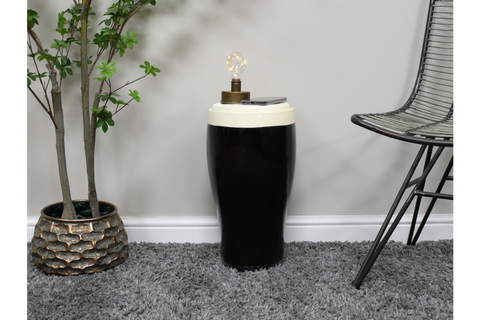 Black and white ceramic stout side table with lamp and phone on top, styled next to chair and plant on gray carpet