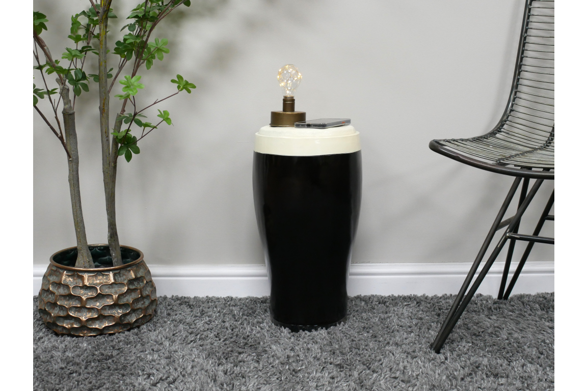 Black and white ceramic stout side table with lamp and phone on top, styled next to chair and plant on gray carpet
