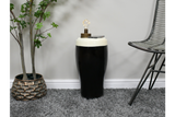 Black and white ceramic stout side table with lamp and phone on top, styled next to chair and plant on gray carpet
