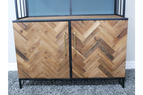 Bar cabinet with herringbone patterned wood doors, black metal frame, gold vertical handles, shown front view on gray carpet.