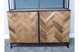 Bar cabinet with herringbone patterned wood doors, black metal frame, gold vertical handles, shown front view on gray carpet.