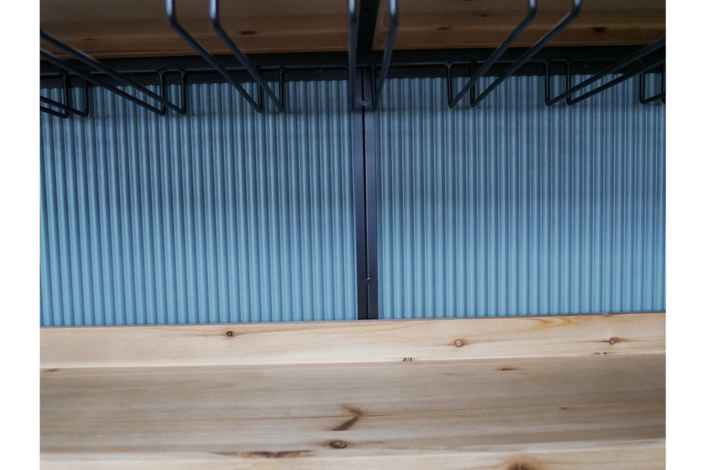 Close-up of light wood bar cabinet shelf with black metal wine glass racks hanging above, blue ribbed background.