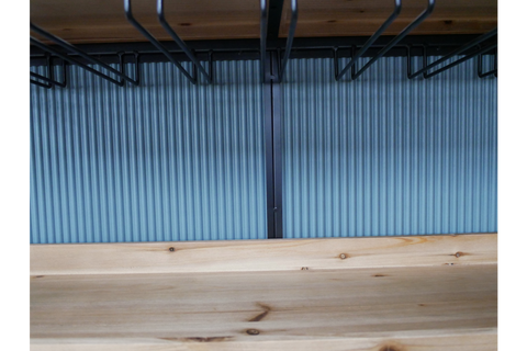 Close-up of light wood bar cabinet shelf with black metal wine glass racks hanging above, blue ribbed background.