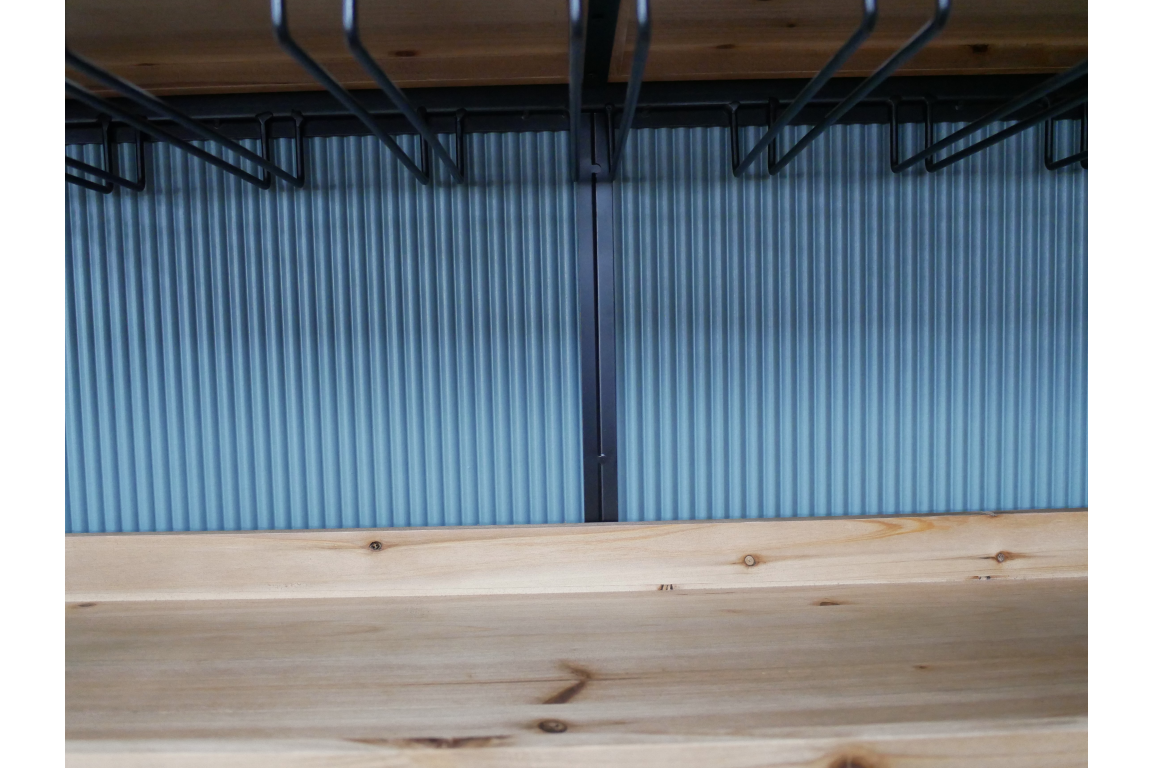Close-up of light wood bar cabinet shelf with black metal wine glass racks hanging above, blue ribbed background.