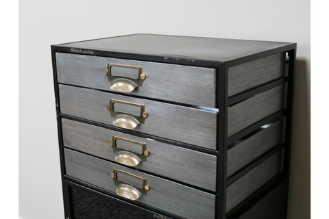 Industrial drawer cabinet with four metal drawers and brass handles in black metal frame, angled view against white wall.