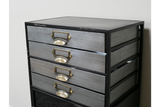 Industrial drawer cabinet with four metal drawers and brass handles in black metal frame, angled view against white wall.