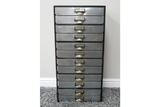 Tall industrial drawer cabinet with ten grey metal drawers and brass handles, standing on grey carpet against white wall.