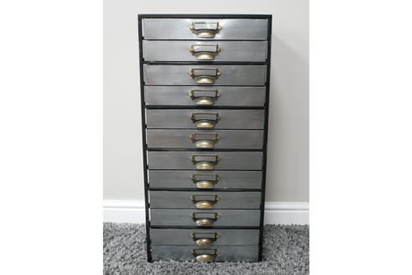 Tall industrial drawer cabinet with ten grey metal drawers and brass handles, standing on grey carpet against white wall.