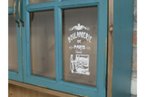 Close-up of distressed blue wooden wall unit with glass pane door featuring vintage white bakery illustration and text.