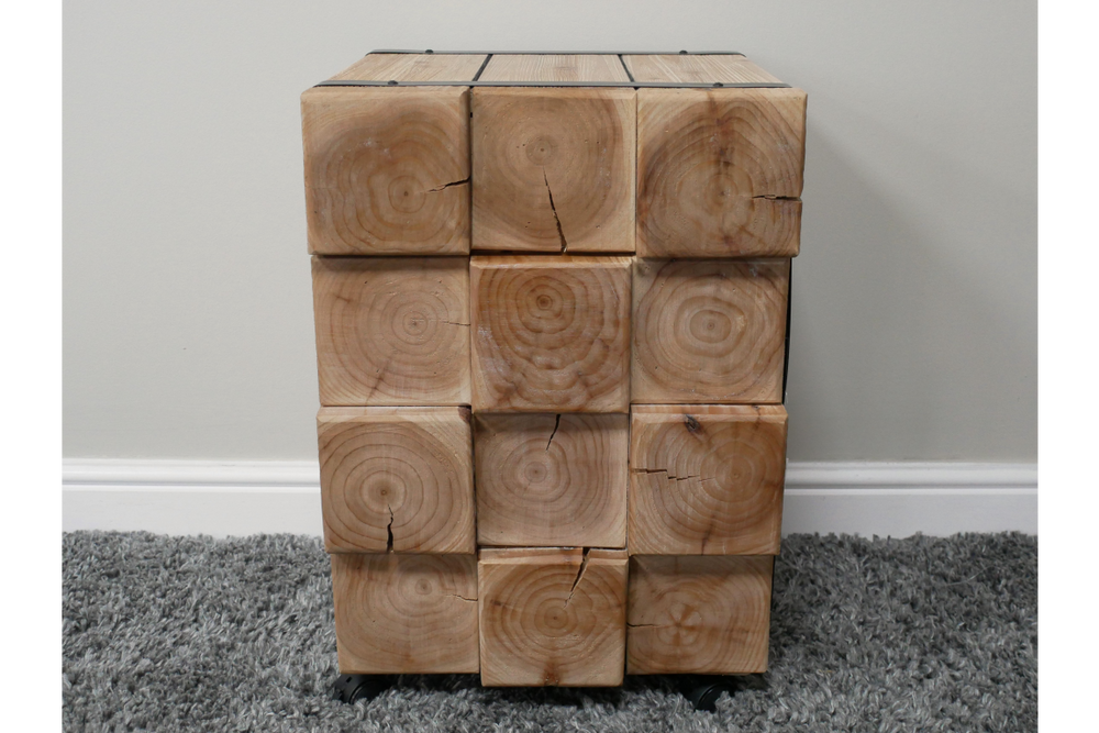 Chunky bedside cabinet made of natural wood blocks with visible grain and cracks, front view on carpeted floor.