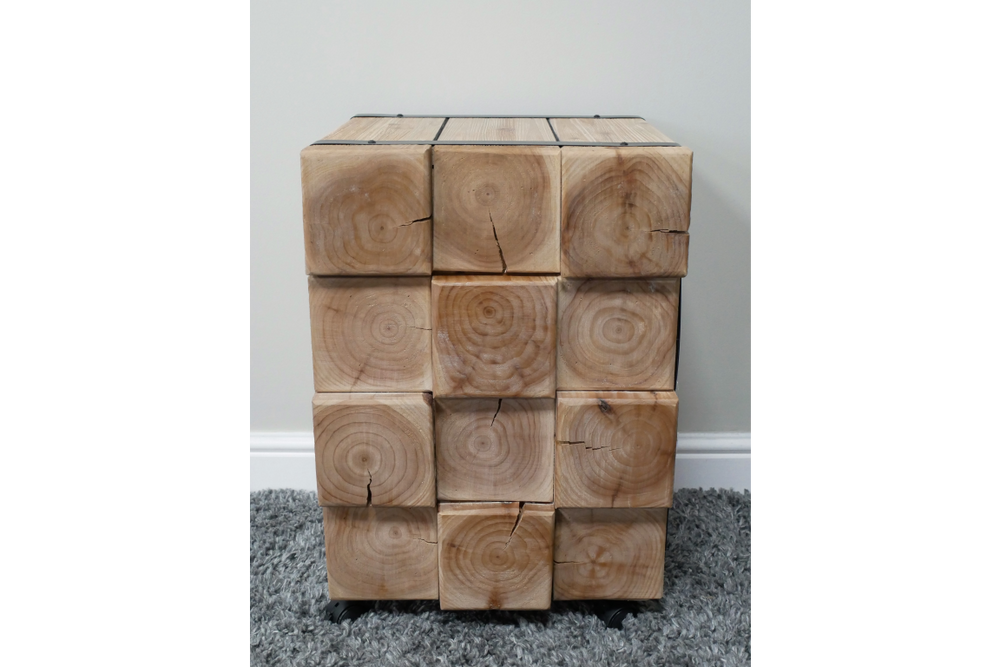Chunky bedside cabinet made of natural wood blocks showing grain and cracks, front view on gray carpet.