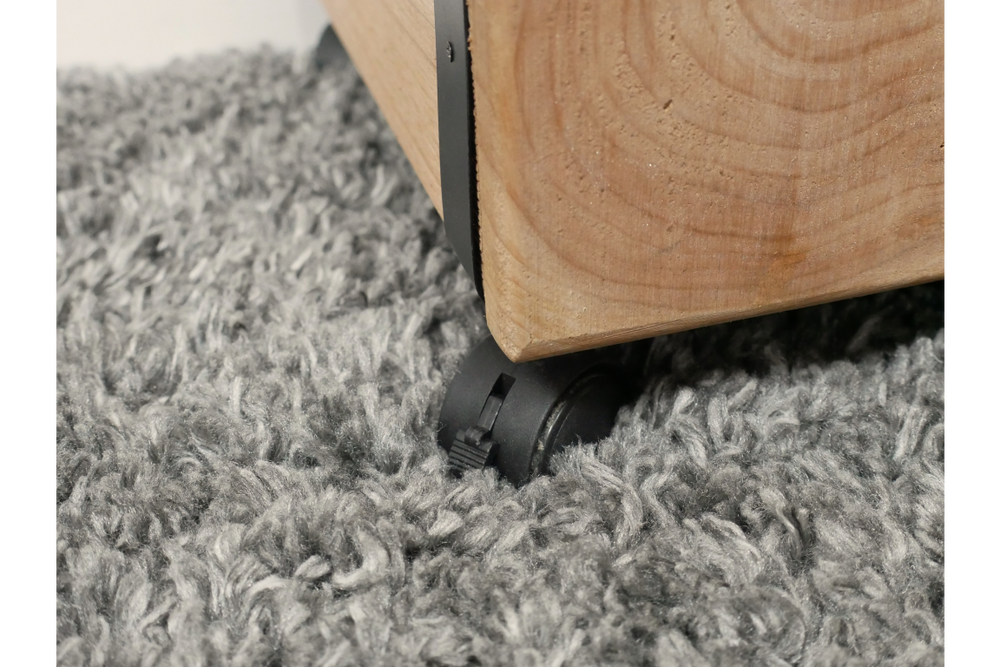 Close-up of light wood chunky bedside cabinet corner with black caster wheel on gray shag carpet