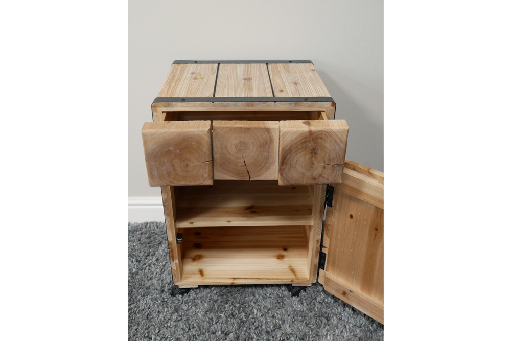 Light wood chunky bedside cabinet with open drawer and door showing interior shelving, placed on gray carpet.