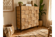 Chunky wooden cabinet with square block front, black metal legs, natural finish, shown in cozy room corner with plants.