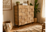 Chunky wooden cabinet with square block front, black metal legs, natural finish, shown in cozy room corner with plants.