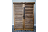 Wooden shutter style bar cabinet with two doors and black vertical handles, photographed front view on carpeted floor.