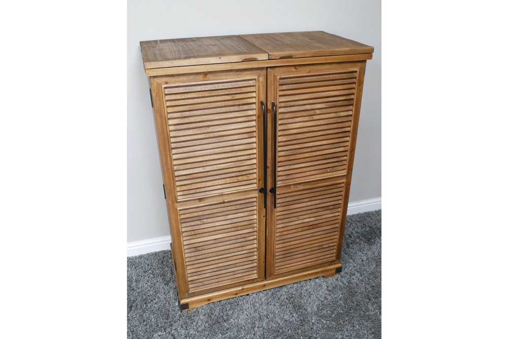 Wooden shutter bar cabinet with two doors, black handles, standing on grey carpet against a white wall.