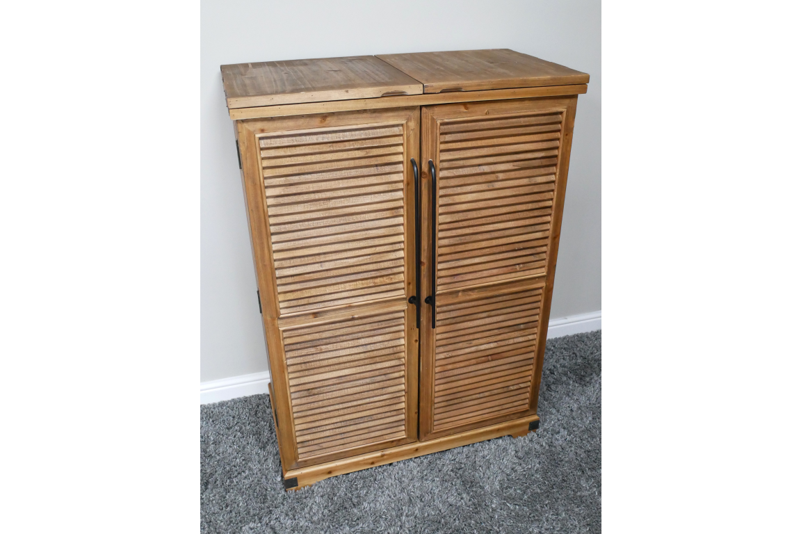 Wooden shutter bar cabinet with two doors, black handles, standing on grey carpet against a white wall.