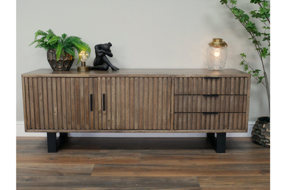 Low wooden TV sideboard with slatted doors and drawers, black metal legs, decorated with plants and lamps, front view.