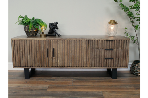 Low wooden TV sideboard with slatted doors and drawers, black metal legs, decorated with plants and lamps, front view.