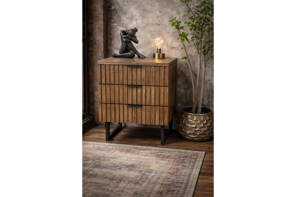 Wooden slatted bedside cabinet with three drawers and black metal legs, shown at an angle in a rustic room setting.