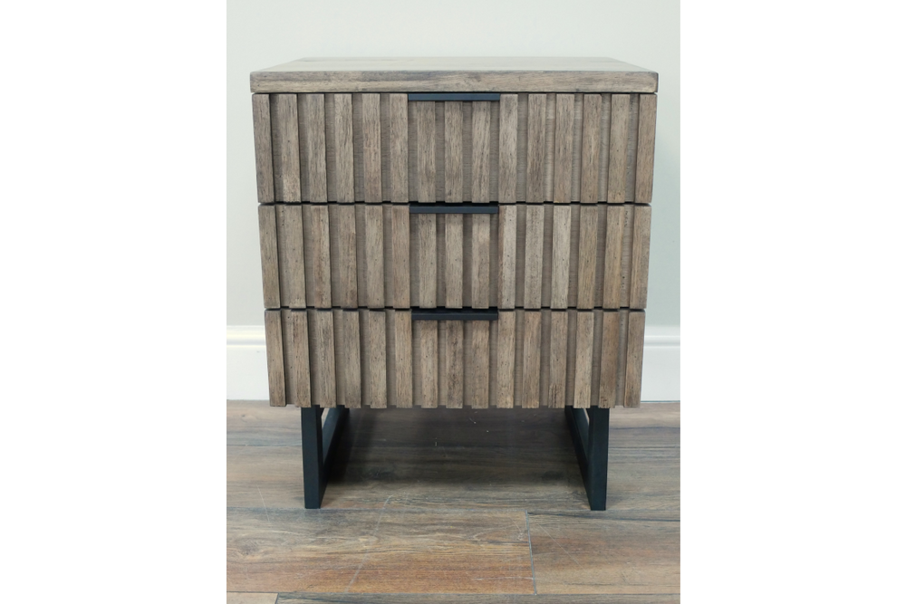 Slatted wooden bedside cabinet with three drawers, black metal handles and legs, front view on wooden floor.