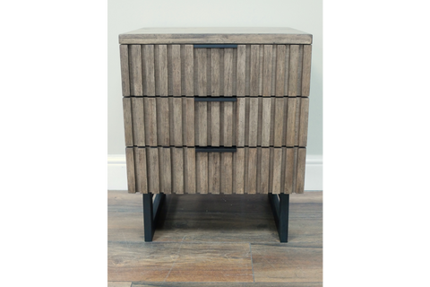 Slatted wooden bedside cabinet with three drawers, black metal handles and legs, front view on wooden floor.