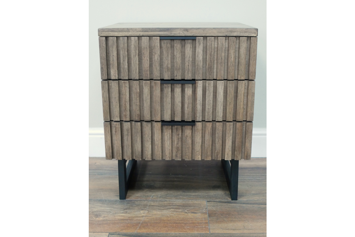 Slatted wooden bedside cabinet with three drawers, black metal handles and legs, front view on wooden floor.