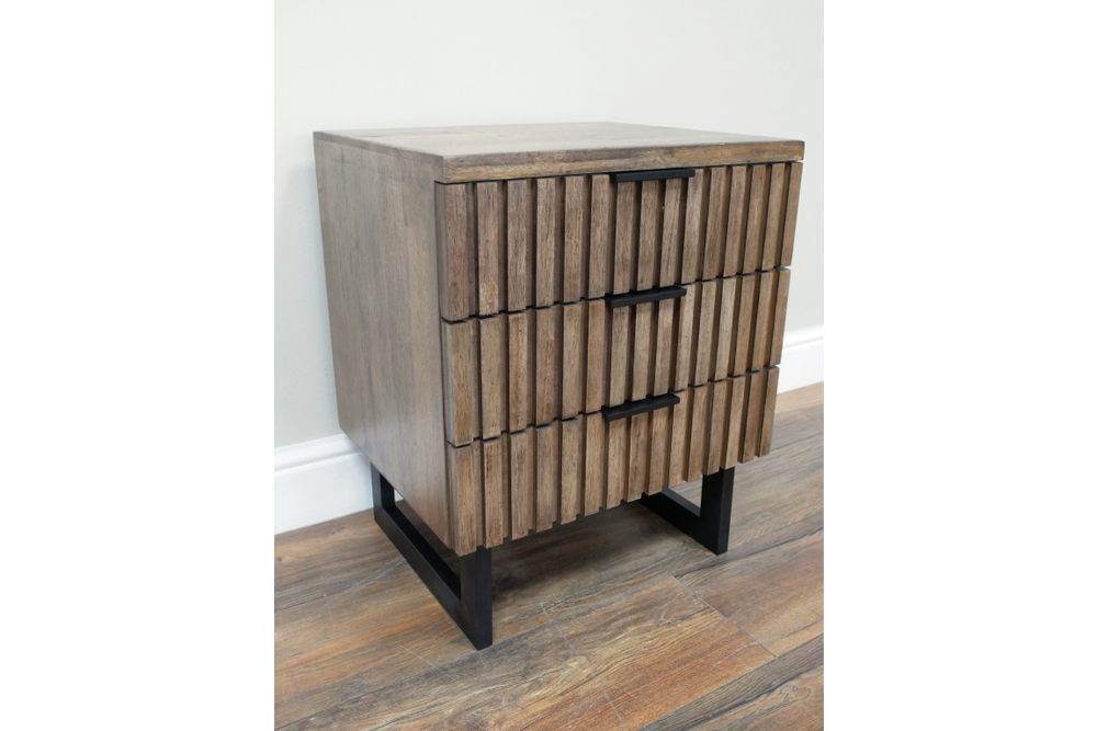 Slatted bedside cabinet in brown wood with three drawers, black metal handles and legs, angled view on wooden floor.