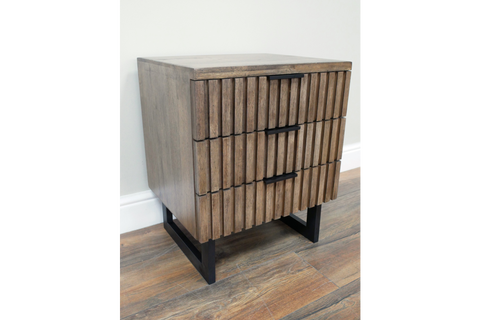 Slatted bedside cabinet in brown wood with three drawers, black metal handles and legs, angled view on wooden floor.