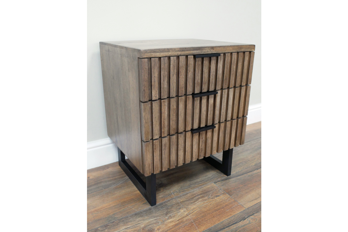 Slatted bedside cabinet in brown wood with three drawers, black metal handles and legs, angled view on wooden floor.