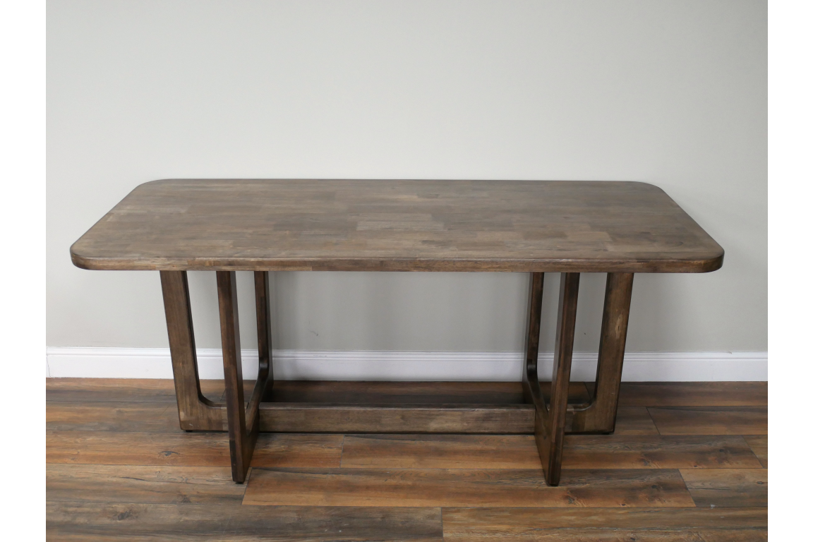 Rectangular wooden dining table with rounded corners and U-shaped legs on wooden floor against light wall.