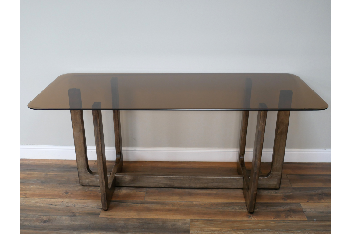 Rectangular dining table with a tinted glass top and wooden trestle legs, shown from front angle on wooden floor.