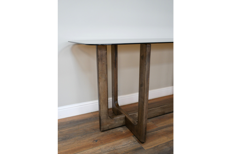 Dining table with clear glass top and wooden legs, side angle view on wooden floor against white wall.