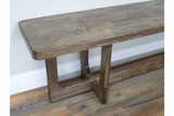 Long wooden bench with sturdy legs, medium brown finish, angled side view on wooden floor against white wall.