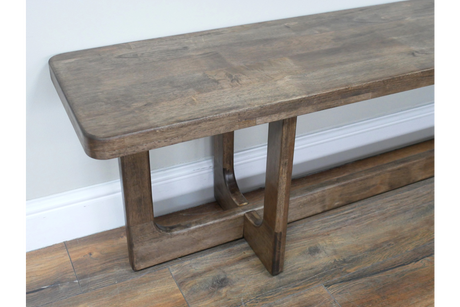 Long wooden bench with sturdy legs, medium brown finish, angled side view on wooden floor against white wall.