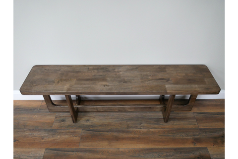 Long wooden bench with thick rectangular top and sturdy legs, placed against light grey wall on wooden floor.