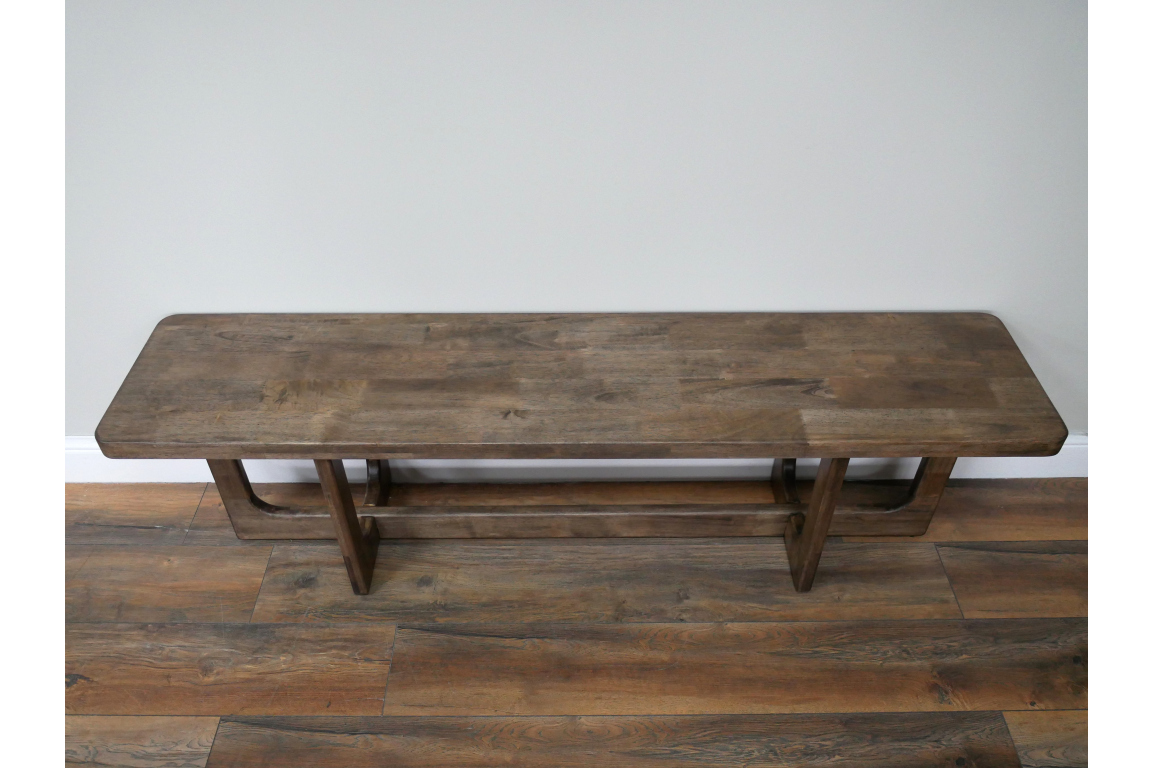 Long wooden bench with thick rectangular top and sturdy legs, placed against light grey wall on wooden floor.