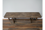 Long wooden bench with thick rectangular top and sturdy legs, placed against light grey wall on wooden floor.