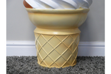 Yellow ceramic side table shaped like an ice cream cone base with a white swirl top, shown from front angle on carpet.