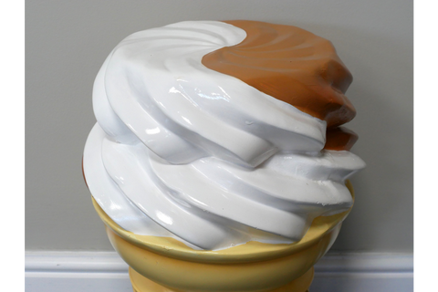 Yellow and white resin side table shaped like a swirl of ice cream with caramel topping, front angle against gray wall.