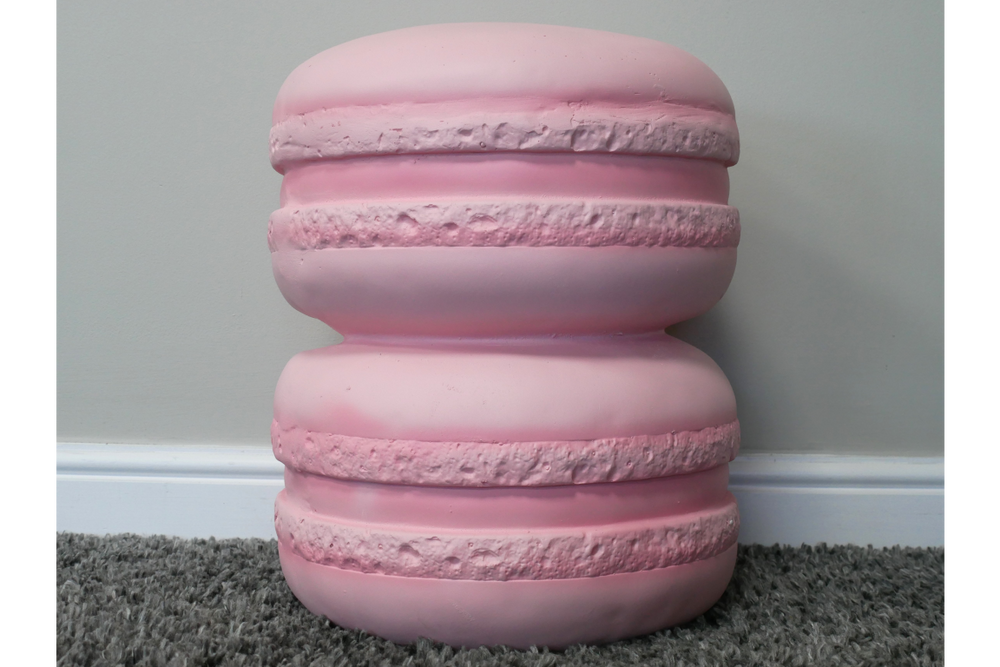 Pink resin side table shaped like two stacked macarons, front view on grey carpet and white baseboard background.
