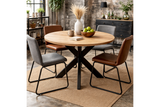 Round dining table with natural wood top and black metal crossed base, shown with mixed grey and brown chairs on woven rug.